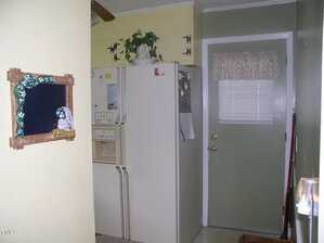 A kitchen corner with a white refrigerator, a green door with a small window and floral curtain, a framed chalkboard on a yellow wall, and a potted plant on top of the fridge.