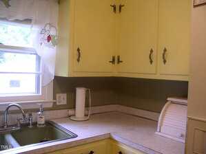 A kitchen countertop with yellow cabinets, a white breadbox, a white electric kettle, a green sink, a soap dispenser, and a window with sheer curtains and a fruit design.