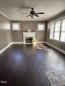 A living room with dark wood floors, brown walls, a white brick fireplace, two small windows above the fireplace, a ceiling fan with light, and a group of windows letting in sunlight on the right.