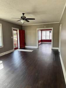 A mostly empty room with dark wood floors, beige walls, and a ceiling fan. A red front door is open to the outside, leading to an attached brightly-lit sunroom with red walls and many windows.