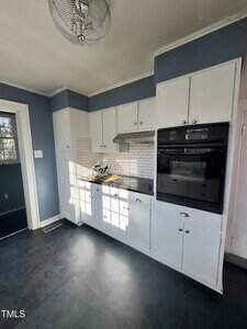 A kitchen with blue walls, white upper and lower cabinets, a white tile backsplash, a black double oven, a gas cooktop with a vent hood, and a black floor. Sunlight streams in through a window.