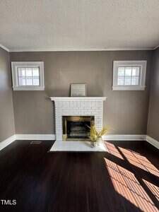 A small living room with dark wood floors, brown walls, and a white brick fireplace centered between two windows. A framed sign and a vase with dried grass sit on the fireplace hearth. Sunlight streams through a window.