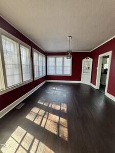 A room with dark wood floors, red walls, multiple large windows with blinds, and a hanging light fixture. Sunlight is streaming through the windows, casting shadows on the floor. An open doorway leads to another room.