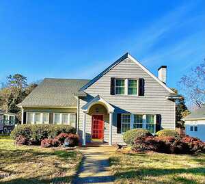 A gray, single-family house at 1922 Oxford Road, Henderson, with black shutters, a steep roof, and a bright red front door. Landscaped bushes adorn the front yard, and a chimney rises on the right side under a clear blue sky.
