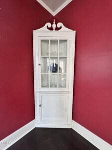 A white, vintage corner cabinet with glass-paneled upper doors and solid lower doors is set against red walls in a room with dark wood floors and white trim.