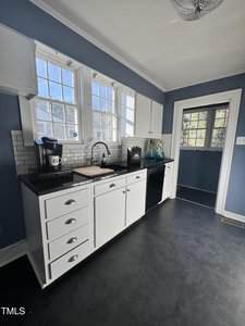 A kitchen with blue walls, white cabinets, black countertops, a black dishwasher, a coffee maker, a sink under double windows, white subway tile backsplash, and a door with a window leading outside.