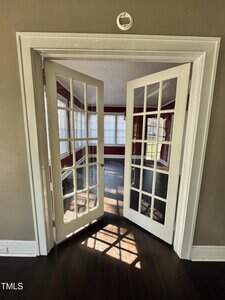 A pair of white French doors open into a sunlit room with large windows, dark wood floors, and red and white walls. Sunlight casts shadows on the floor through the glass panes of the doors.