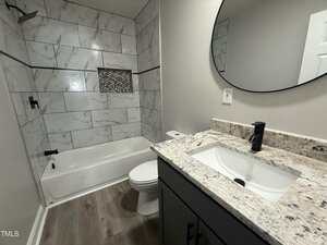 Modern bathroom with a white bathtub and shower, gray marble tile surround, built-in shower niche, granite countertop vanity with under-mount sink, round mirror, and wood-look flooring.