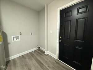 A small laundry room with light gray walls, wood-look flooring, exposed utility hookups, a black door with gold hardware, and a corner of a water heater visible on the left side.
