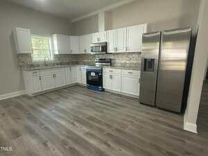 Modern kitchen with white cabinets, stainless steel appliances including a refrigerator, oven, and microwave, marble-patterned backsplash, and wood-look flooring. A window is above the sink on the left side.
