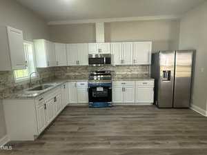 A modern kitchen with white cabinets, marble countertops, a stainless steel stove and microwave, a double-door refrigerator, a single sink, and light wood flooring. A window is above the sink on the left wall.