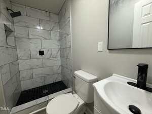 A modern bathroom with a walk-in shower featuring white marble-style tiles and a black showerhead, a white toilet, and a white sink with a black faucet beneath a mirror.