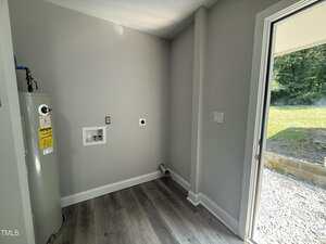 A small laundry room with light gray walls, a water heater on the left, hookups for a washer and dryer, laminate flooring, and a door leading outside to a grassy yard.