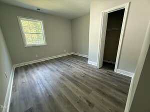 An empty room with light gray walls, a window letting in natural light, a small open closet, white baseboards, and dark wood-style flooring.