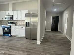 A modern kitchen with white cabinets, stainless steel appliances, marble backsplash, and light wood flooring opens into a hallway leading to a closed black door.