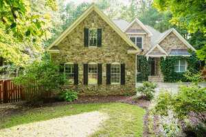 A two-story house with a stone exterior, black shutters, and multiple windows. The front yard has green shrubs, trees, and a wooden fence to the left, with a driveway on the right side.