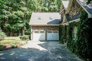 A two-car garage with white doors is attached to a house with stone siding and ivy climbing up the exterior walls. A concrete driveway and landscaped yard with trees are visible.