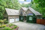A two-story house with a stone exterior at 65 Hidden Cove Court, Youngsville, features ivy climbing the front, three white garage doors, and a paved driveway, all surrounded by trees and greenery.