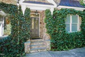A stone house entrance with a gray door featuring decorative glass panels, two lantern-style wall lights, and ivy growing around the doorway and windows. Stone steps lead up to the front door.