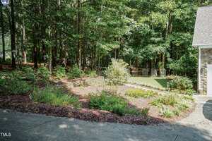 A landscaped garden bed with mulch and various small shrubs and plants sits next to a concrete driveway, bordered by tall trees in a wooded area.