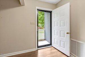 A white door with a gold knob is open, revealing a screen door and a view of a small outdoor patio with greenery. The room has beige walls, wood flooring, and a vent near the door.
