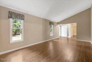 A spacious, empty living room with light beige walls, wood flooring, two windows with valances on the left, and an open doorway leading to a white front door. Natural light enters from the windows and door.