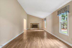 A spacious, empty living room with light-colored walls, vaulted ceiling, wood flooring, a large window with a valance, and a stone fireplace centered on the far wall.