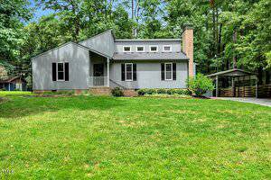 A gray, single-story house at 703 Forrest Lane in Creedmoor features a small front porch, multiple windows, a brick chimney, and an adjacent carport, all set on a grassy lawn with trees in the background.