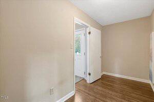 A small, empty room with beige walls, white trim, and wood flooring. There is a closed white door and an open doorway leading to another area with a white exterior door and a window.