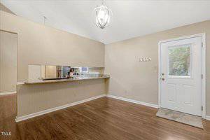 A room with beige walls, wood flooring, a granite countertop pass-through to the kitchen, a white door with a window, a wall hook rack, and a ceiling light fixture. A small mat is placed by the door.