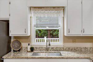 A kitchen countertop with a stainless steel sink under a window with blinds and a white valance. White cabinets are above and below the counter, and decor items sit next to the sink. Trees are visible outside the window.