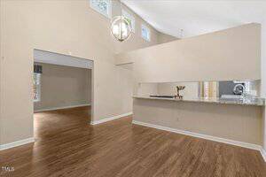 A spacious room with light beige walls, wood flooring, high ceilings, a modern chandelier, and a long granite countertop that opens to another room. Multiple small windows provide natural light.