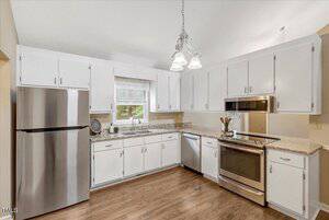 A kitchen with white cabinets, stainless steel appliances including a refrigerator, dishwasher, and oven, granite countertops, a window over the sink, and wood flooring.