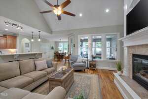 A spacious living room with a beige sectional sofa, armchair, coffee table, and patterned rug. The room has hardwood floors, a ceiling fan, fireplace, large windows, and an open view of the kitchen and dining area.