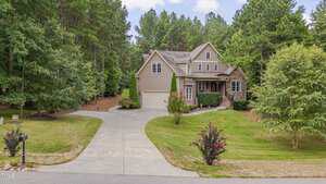 A two-story house with tan siding and a three-car garage sits at the end of a wide, curved driveway. The property is surrounded by green grass, trees, and shrubs under a partly cloudy sky.