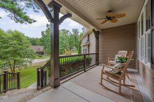 A covered front porch with two wooden rocking chairs, a ceiling fan, potted flowers, and a rug, overlooking a green lawn and a suburban neighborhood street.