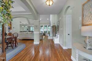 A bright entryway with hardwood floors leads to an open living and dining area. The space features neutral walls, recessed lighting, arched doorways, and large windows providing natural light.