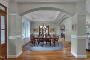 A dining room with a wooden table and eight chairs on a round braided rug, a chandelier overhead, and a glass cabinet against the back wall. The room opens to a kitchen and living area with hardwood floors throughout.
