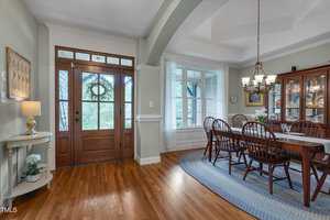 A dining room with wood floors, a large window, glass-front china cabinet, oval rug, wooden dining table with six chairs, and a front door with glass panels and a wreath. A small shelf with a plant is near the entry.