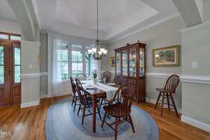 A formal dining room features a wooden table with six chairs, a patterned rug, a chandelier, a glass-front china cabinet, a framed painting on the wall, and large windows letting in natural light.
