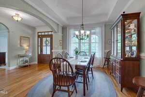 A dining room with a rectangular table, six wooden chairs, a chandelier, a china cabinet, hardwood floors, and large windows letting in natural light. A blue rug is under the table and a front door is visible.