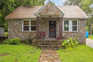 A small stone house with a gable roof at 630 East Avenue, Henderson, features a front porch, stone steps, and lush green grass. Two signs are displayed in the front windows, and a chair sits on the porch amid mature trees.
