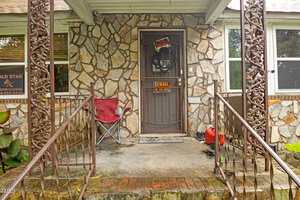 A stone porch with a metal railing, a red folding chair, a black screen door displaying a "BEWARE OF DOG" sign, and a small red gas can on the ground. A window and business sign are visible on the left.