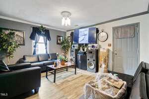 A living room with black leather sofas, a coffee table, large speakers, a wall-mounted TV, and potted plants. A clock hangs near a closed gray door. Grocery bags and boxes sit in the corner by the door.