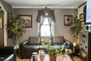 A living room with dark leather couches, a central window with dark curtains, framed art on the walls, potted artificial plants, a coffee table with various items, and a large speaker in the right corner.