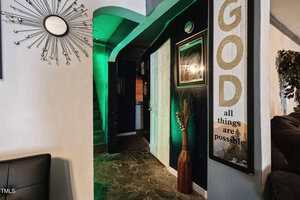 A hallway with dark walls features a large framed sign reading "GOD all things are possible," a decorative vase with dried plants, a sunburst wall clock, and a partially visible living area. Marble-patterned flooring is present.