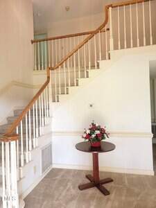 A small wooden round table with a floral arrangement sits below a carpeted staircase with white spindles and a wooden handrail in a bright, neutral-toned entryway.