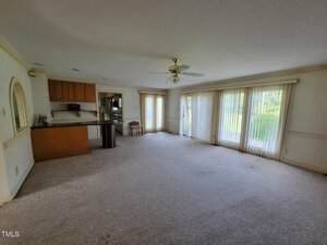 Spacious living area with carpeted floor, ceiling fan, and large windows with vertical blinds. A kitchen with bar seating is visible in the background. Natural light enters from the windows and sliding glass door.