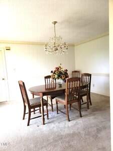 A dining room with a wooden table, six matching chairs, a vase of colorful artificial flowers as a centerpiece, a crystal chandelier above, and light-colored carpeted flooring. The walls are light with decorative trim.