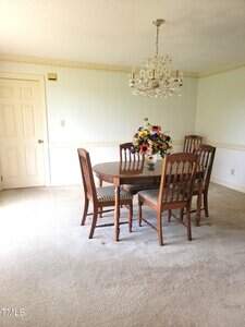 A dining room with a round wooden table and four matching chairs, a vase of colorful flowers on the table, a chandelier overhead, and a closed door in the background. The room has carpeted floors and white walls.